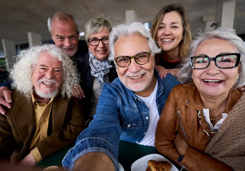 Happy mature gray hair man taking selfie of group seniors Caucasian cheerful friends posing together sitting at cafeteria. Older people looking smiling at camera with snacks on table nursing home